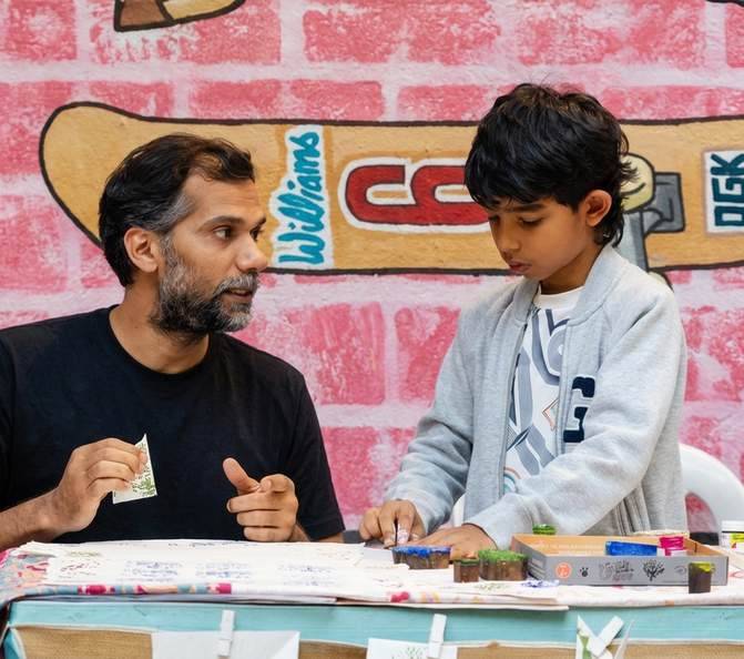 An artist and a boy painting together at a table in front of a mural
