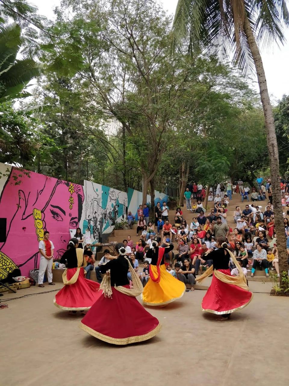 Dancers in swirling red and yellow skirts performing on the lower landing, with neighbours seated on the steps beneath the trees